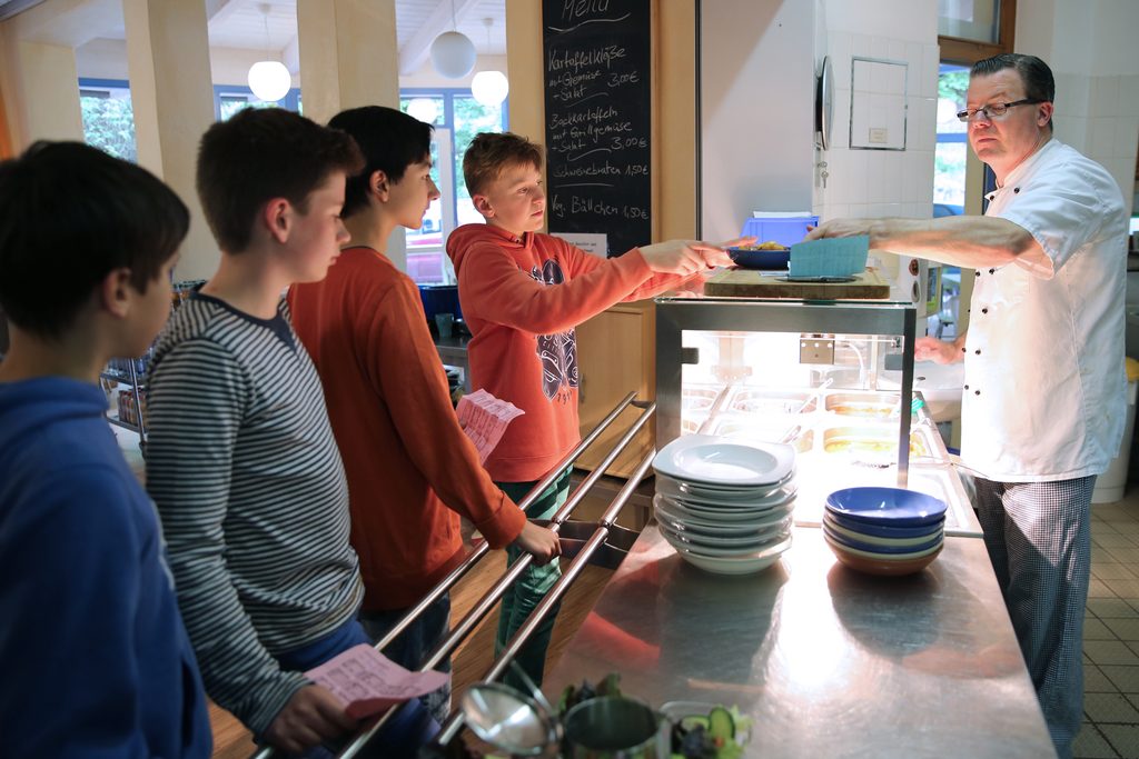 4 Schüler stehen vor der Essensausgabe-Theke in der Kantine und bekommen von dem Koch einen Teller mit einer Mahlzeit.