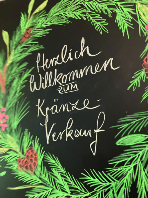 Auf eine Tafel ist ein grüner Adventskranz gemalt und die Aufschrift