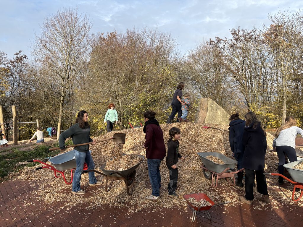 Eltern und Kinder stehen mit Schubkarren und Schaufeln auf dem Schulgelände vor einem Haufen Hackschnitzel.
