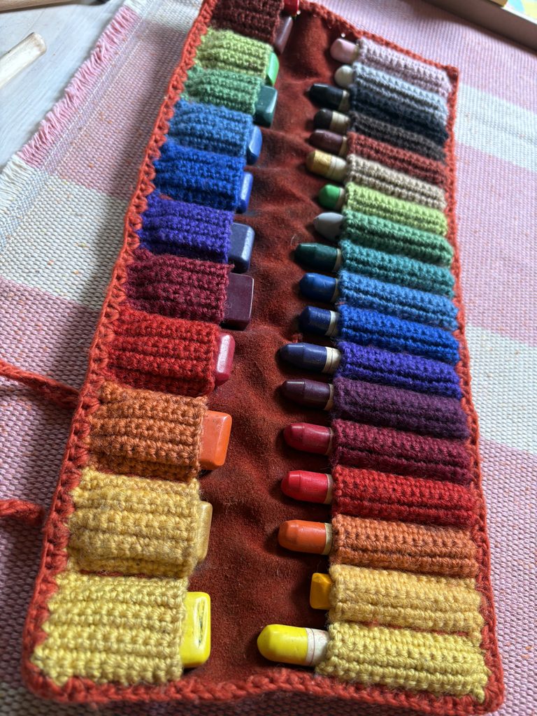 Ein in Regenbogen gehäkeltes Rollmäppchen mit den farblich dazu passenden Wachsmalblöckchen und Stiften.