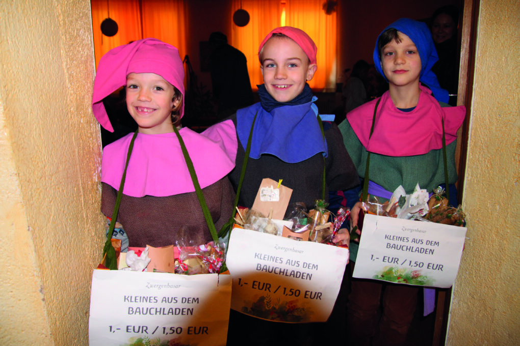 3 Kinder sind als Wichtel verkleidet, stehen nebeneinander und haben Bauchläden mit Plätzchen und anderen Leckereien darin, zum Verkauf.